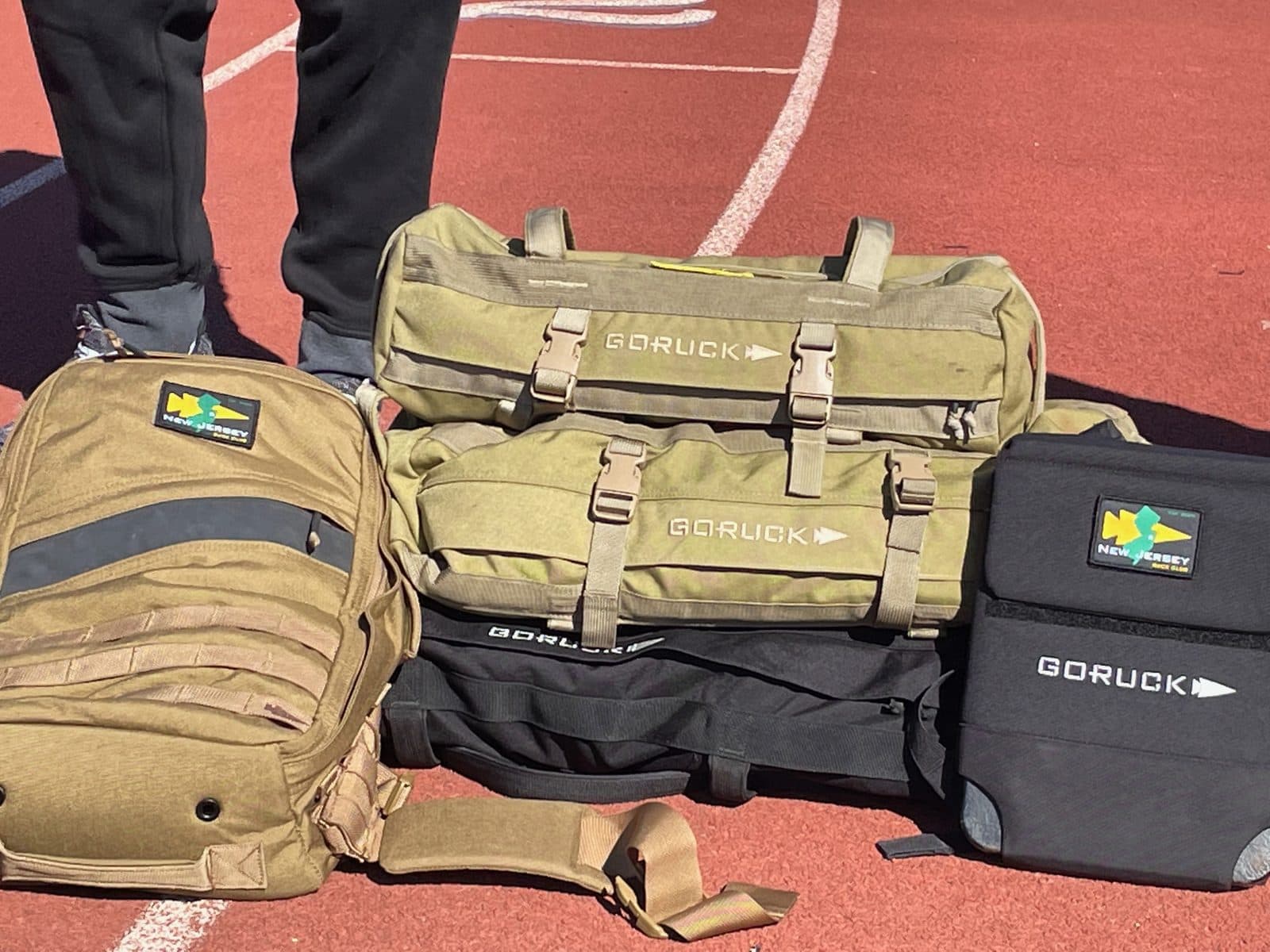 Rucksacks and sandbags with New Jersey Ruck Club patches lined up on a running track